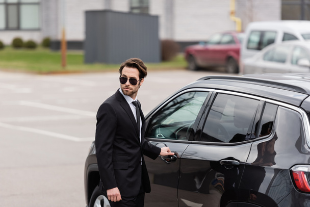 bodyguard in suit and sunglasses with security earpiece opening door of modern auto  - Photo, Image