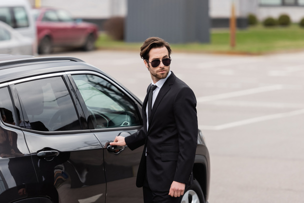bodyguard in sunglasses with security earpiece opening door of modern auto  - Foto, afbeelding