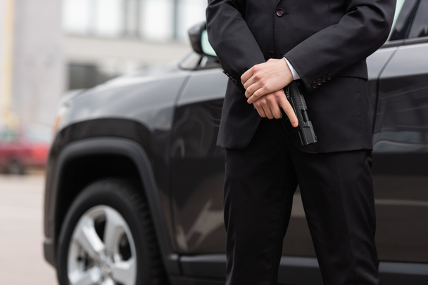 cropped view of bodyguard in suit holding gun near blurred modern auto - Foto, afbeelding