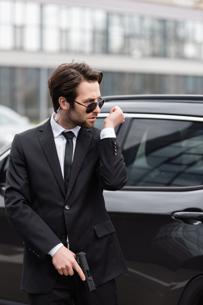 bodyguard in sunglasses with security earpiece holding gun and talking near modern auto  - Foto, Imagem