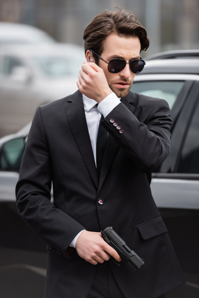 szakállas testőr napszemüvegben, biztonsági fülhallgatóval a kezében fegyver és beszél közel modern autó  - Fotó, kép