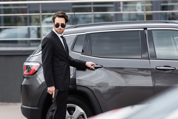 safeguard in sunglasses and suit holding gun near modern auto - Zdjęcie, obraz