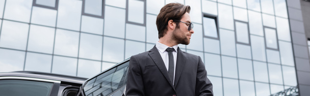 side view of bearded bodyguard in suit near car and modern building, banner - Photo, Image