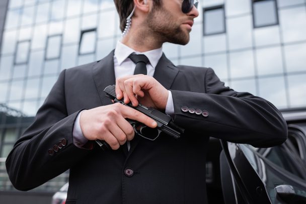 side view of bearded bodyguard in suit holding gun near modern building  - Photo, Image