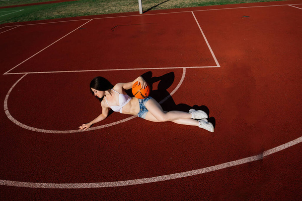 若い運動選手の女性は、トップとスウェットパンツで、屋外のバスケットボールコートでボールと遊ぶ. - 写真・画像