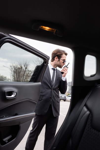 bodyguard in sunglasses using walkie talkie near modern car - Zdjęcie, obraz