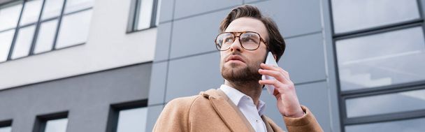 vista basso angolo di uomo d'affari barbuto in occhiali e cappotto beige parlando sul cellulare vicino edificio esterno, banner - Foto, immagini