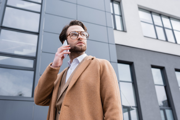 matala kulma näkymä miehen lasit ja beige takki puhuu matkapuhelin lähellä rakennuksen ulkopuolella  - Valokuva, kuva