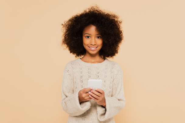 Gülümseyen Afrikalı Amerikalı genç kız ellerinde akıllı telefonla bejde izole bir şekilde bekliyor. - Fotoğraf, Görsel