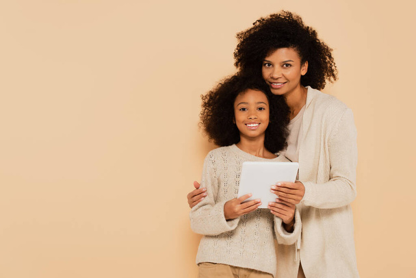 african american adult mother hugging smiling preteen daughter with tablet in hands isolated on beige - Fotó, kép