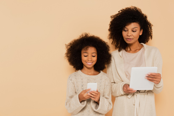 smiling african american preteen daughter and adult mother holding cellphone and tablet in hands isolated on beige - Fotoğraf, Görsel