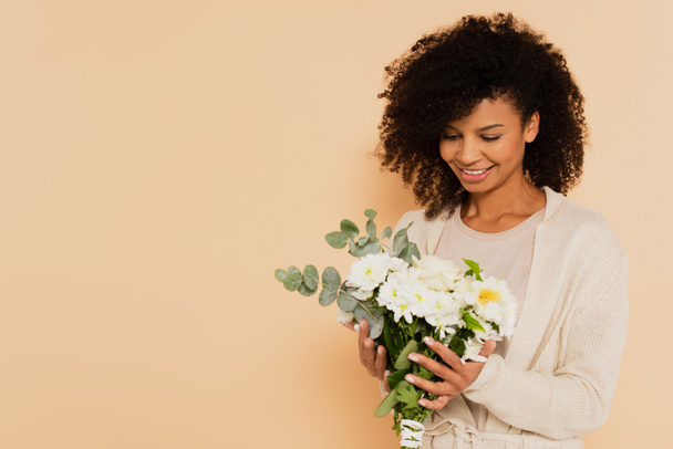 afro-américaine tenant bouquet de marguerites et souriant sur fond beige - Photo, image