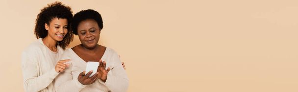 Afro-Amerikan yetişkin kızı, bej arka planda, pankartta orta yaşlı bir annenin ellerinde, parmağıyla akıllı telefonu işaret ediyor. - Fotoğraf, Görsel