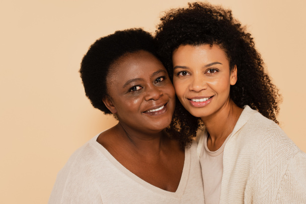 sorridente afro-americano adulto filha e meia-idade mãe abraçando bochecha a bochecha isolado no bege - Foto, Imagem
