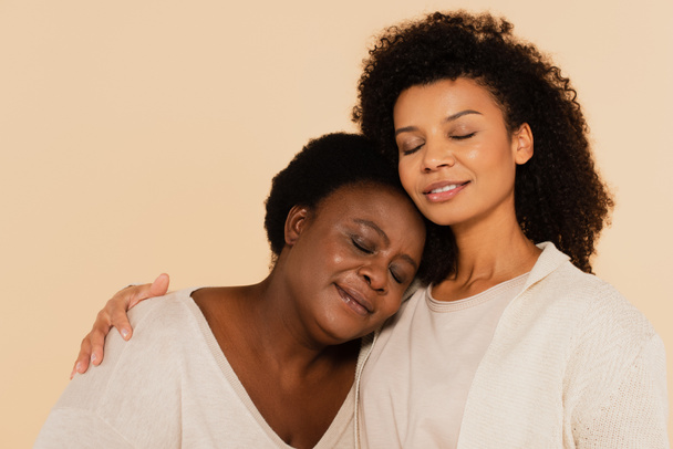 calma afroamericana hija adulta y madre de mediana edad abrazo con los ojos cerrados aislados en beige - Foto, Imagen