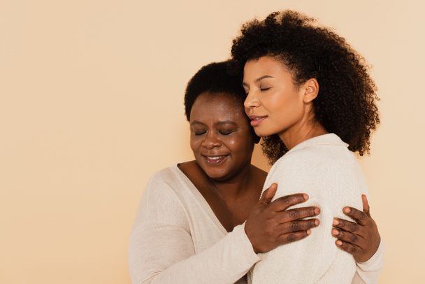 portrait d'une fille adulte afro-américaine et d'une mère d'âge moyen embrassant les yeux fermés isolés sur du beige - Photo, image