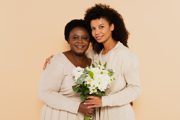 Afrikalı Amerikalı yetişkin kız ve orta yaşlı anne bej rengi bir buket çiçekle dikiliyorlar. - Fotoğraf, Görsel