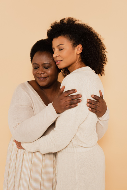 african american adult daughter and middle aged mother hugging with closed eyes isolated on beige - Photo, Image