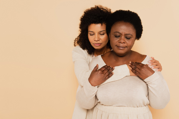 african american middle aged mother and adult daughter hugging and holding hands with closed eyes isolated on beige - Photo, Image