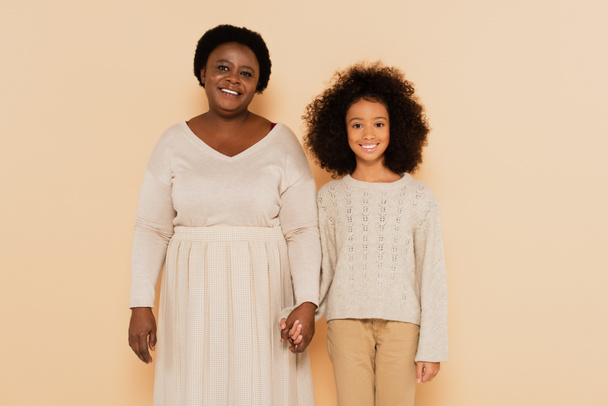 afro-américaine grand-mère et petite-fille tenant la main dans des vêtements pastel sur fond beige - Photo, image