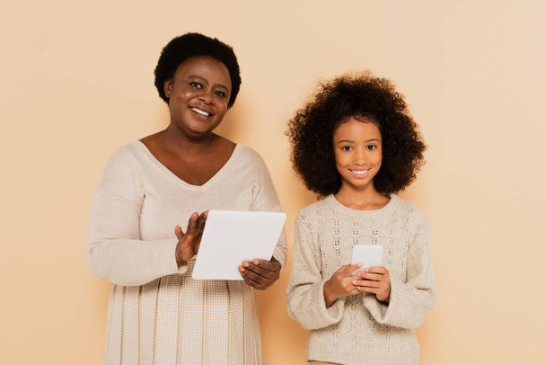 african american granddaughter with cellphone and grandmother with tablet looking at camera on beige background - Foto, Imagem