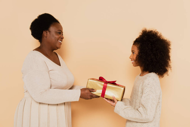 oldalnézetben mosolygós afro-amerikai unoka és nagymama holding ajándék doboz bézs háttér - Fotó, kép