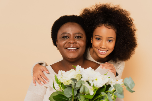 happy african american grandmother and granddaughter with bouquet of flowers isolated on beige - Zdjęcie, obraz