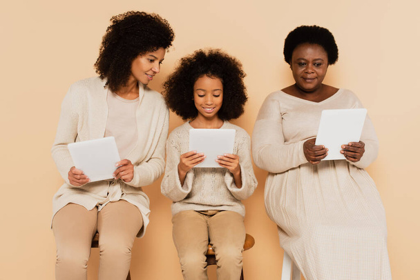 african american daughter, granddaughter and grandmother sitting and looking at tablets on beige background - Photo, Image