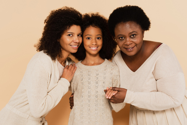 Afrikaans amerikaanse dochter, kleindochter en grootmoeder knuffelen en kijken naar camera op beige achtergrond - Foto, afbeelding