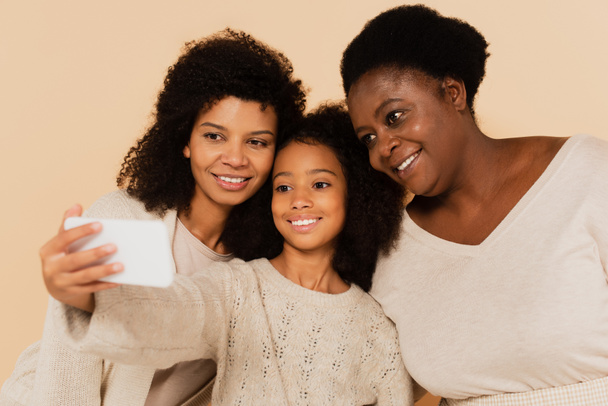 smiling african american daughter, granddaughter and grandmother taking selfie on cellphone together on beige background - Photo, Image
