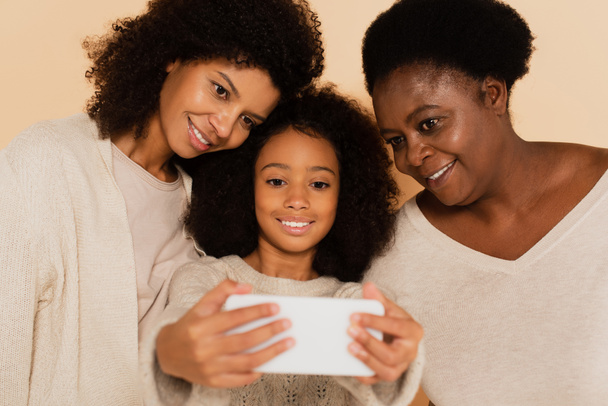 african american daughter, granddaughter and grandmother taking selfie on cellphone together on beige background - Foto, afbeelding