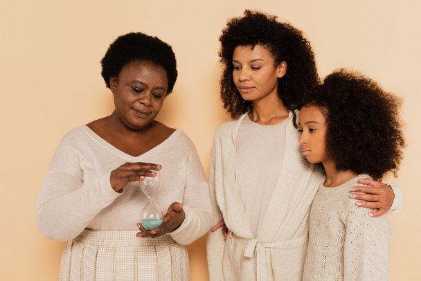 アフリカ系アメリカ人の祖母が砂時計を持っていて娘と孫娘がベージュの背景に立っています - 写真・画像