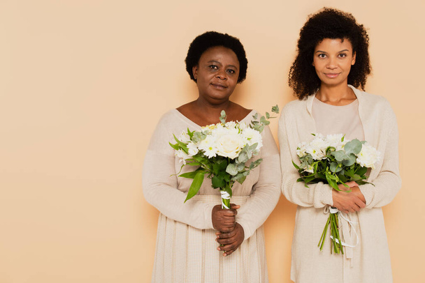 africano americano de mediana edad y mujeres adultas mirando a la cámara con ramos de flores sobre fondo beige - Foto, Imagen