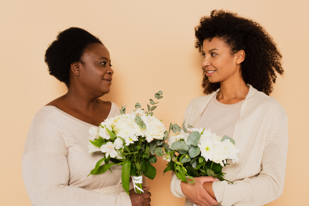 afrikanisch-amerikanische Mutter mittleren Alters und erwachsene Tochter betrachten einander mit Blumensträußen auf beigem Hintergrund - Foto, Bild