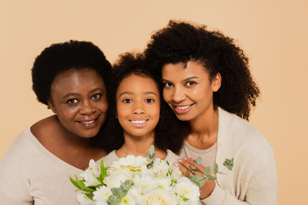 happy african american family of mother, daughter and grandmother hugging and holding bouquet of flowers isolated on beige - Photo, Image