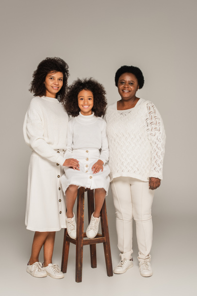 happy african american mother and grandmother holding hands with granddaughter sitting on stool on grey background - Foto, afbeelding