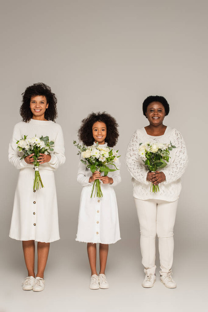 volledige weergave van Afrikaanse Amerikaanse moeder en grootmoeder op zoek naar kleindochter en het houden van boeketten van bloemen op grijze achtergrond - Foto, afbeelding