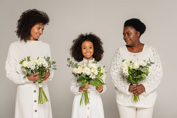 アフリカ系アメリカ人の母親と祖母が孫娘を見て灰色で隔絶された花の花束を持っています - 写真・画像