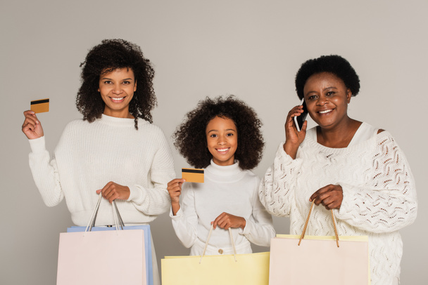 madre e hija afroamericana sosteniendo tarjetas de crédito y bolsas de compras cerca de la abuela hablando por teléfono aislado en gris - Foto, Imagen