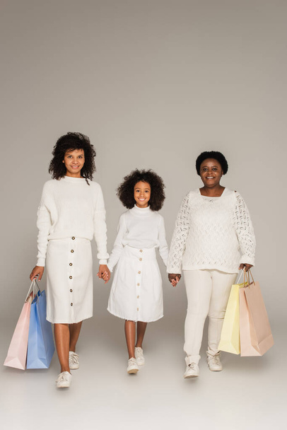 Αφροαμερικανή μητέρα, κόρη και γιαγιά περπατούν με τσάντες για ψώνια και κρατιούνται χέρι-χέρι σε γκρι φόντο - Φωτογραφία, εικόνα