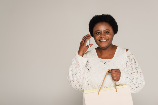 smiling african american middle aged woman holding shopping bags and speaking on cellphone isolated on grey - Foto, Bild