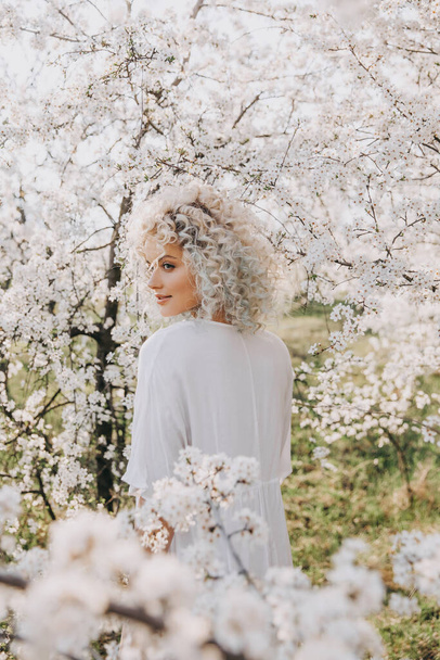 Young blonde woman with curly hair, wearing a white dress, posing next to a cherry tree in bloom. - Foto, Imagen