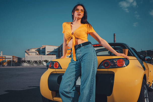 Jonge brunette vrouw in blauwe jeans, oranje top en zonnebril poseert in de buurt van gele auto roadster met papieren kopje thee op de kofferbak. Zomer zonnige dag op de parkeerplaats. Sluiten, kopieerruimte, modelleren - Foto, afbeelding