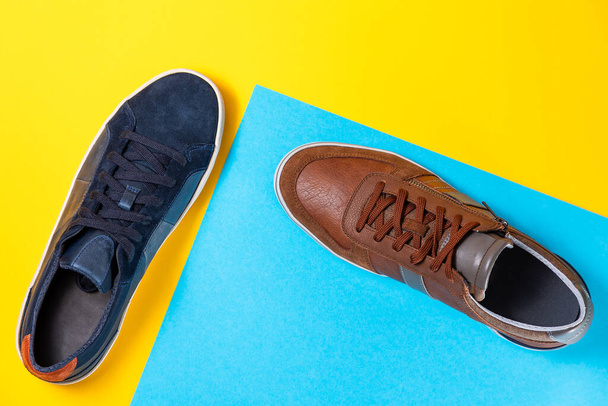 Δύο ανδρικά stylish sneakers από μπλε και καφέ δέρμα σε κίτρινο-μπλε φόντο. Επίπεδη. Αντιγραφή χώρου. Έννοια των αγορών και των πωλήσεων. - Φωτογραφία, εικόνα