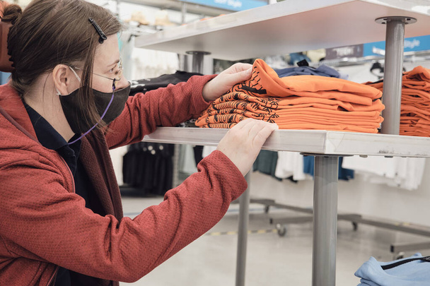 Жінка в масці проти вірусу вибирає помаранчеві сорочки в магазині одягу, щоб купити
 - Фото, зображення