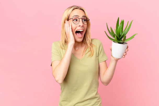 young blonde woman feeling happy, excited and surprised and holding a cactus - Foto, Imagen