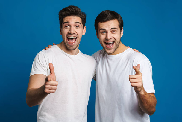 Excited handsome guys screaming and pointing finger at camera isolated over blue background - Фото, изображение