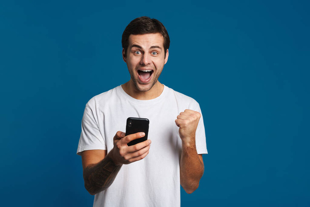 Delighted guy making winner gesture while using mobile phone isolated over blue background - Photo, Image