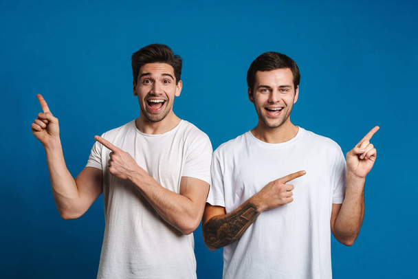Excited unshaven two guys smiling while pointing fingers at opposite sides isolated over blue background - Fotografie, Obrázek