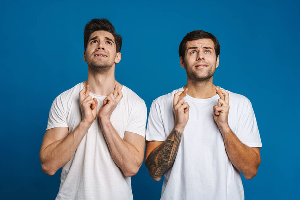 Tense handsome guys posing with fingers crossed for good luck isolated over blue background - 写真・画像